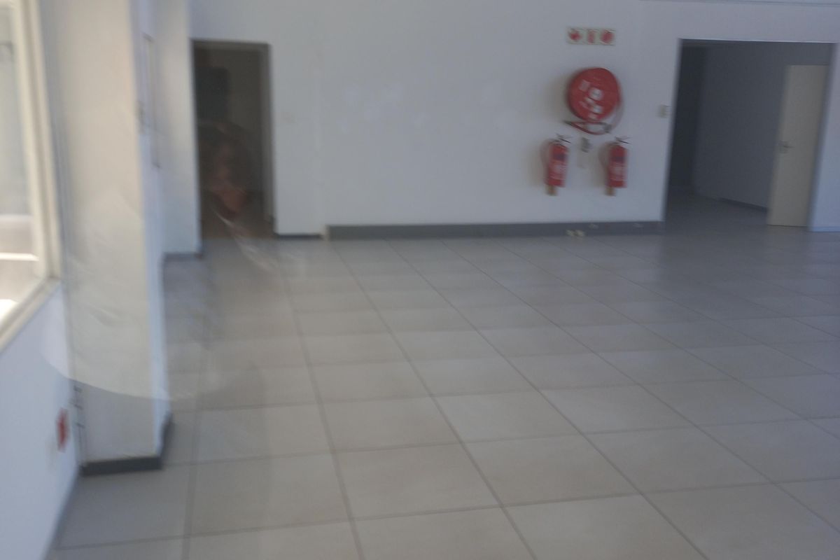 A spacious, empty office area featuring tiled flooring, a suspended ceiling, and wall-mounted fire safety equipment.