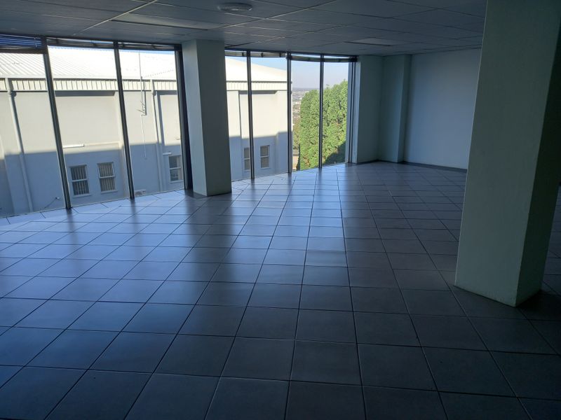 An empty, open-plan office space featuring large floor-to-ceiling windows, tiled flooring, and a suspended ceiling, offering ample natural light and a professional layout.