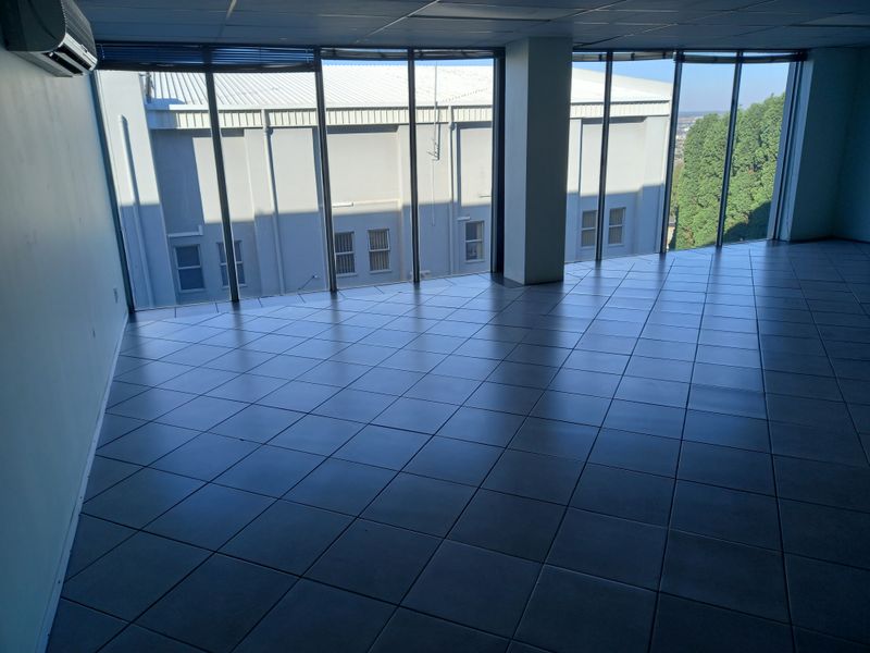 A bright, empty office space featuring large floor-to-ceiling windows, tiled flooring, and a wall-mounted air conditioning unit.