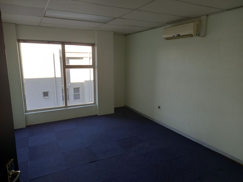 An empty office space featuring a suspended ceiling, split-system air conditioning unit, and blue carpet tiles with a large window providing natural light.