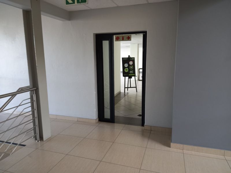 A clean, neutral-toned office hallway featuring tiled flooring and a metal balustrade, leading into an adjacent office space.