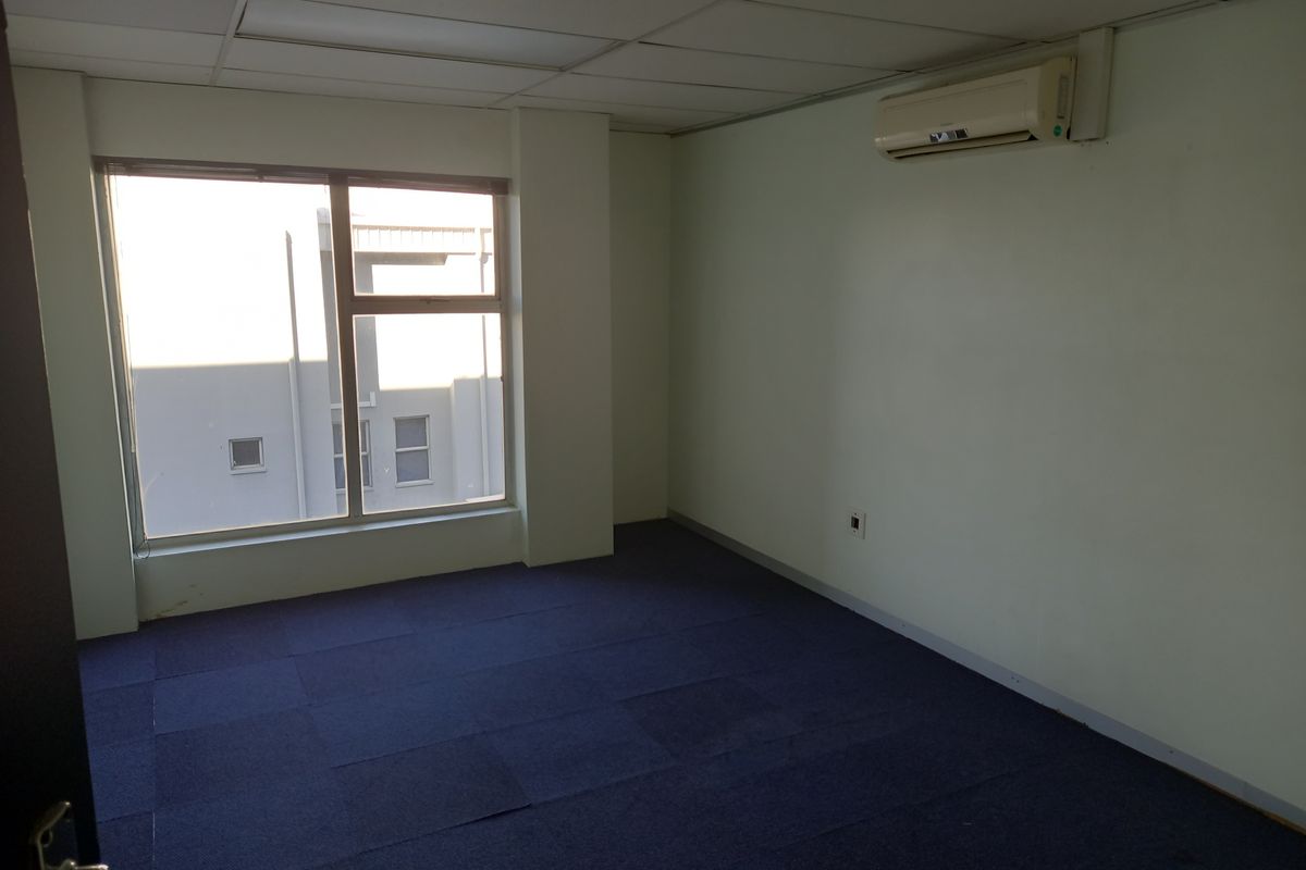 An empty office space featuring a suspended ceiling, split-system air conditioning unit, and blue carpet tiles with a large window providing natural light.