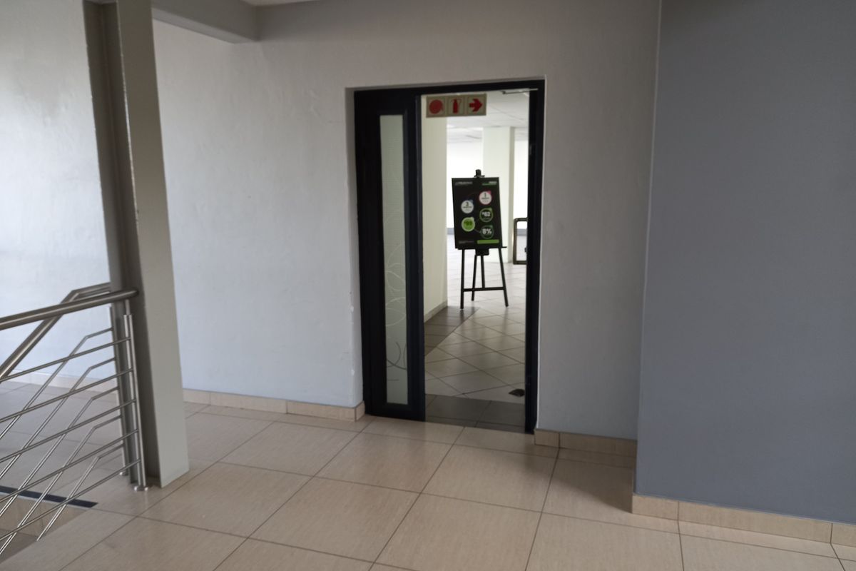 A clean, neutral-toned office hallway featuring tiled flooring and a metal balustrade, leading into an adjacent office space.
