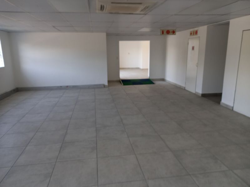 An empty, open-plan office space featuring tiled flooring, a suspended ceiling with integrated air conditioning, and clear access to adjacent rooms.