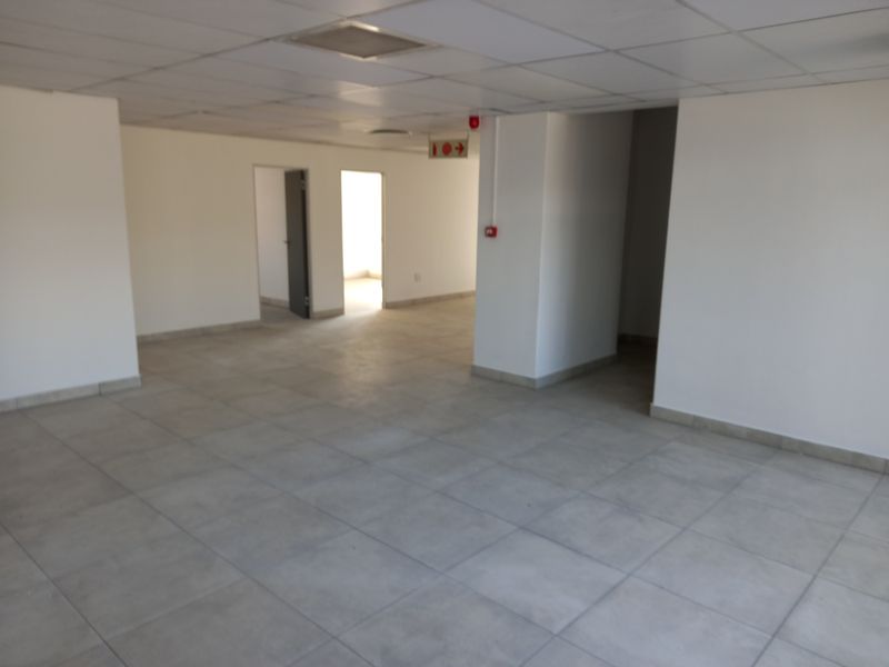 An open-plan office space featuring neutral ceramic floor tiles, a suspended ceiling, and clear access to adjacent rooms.