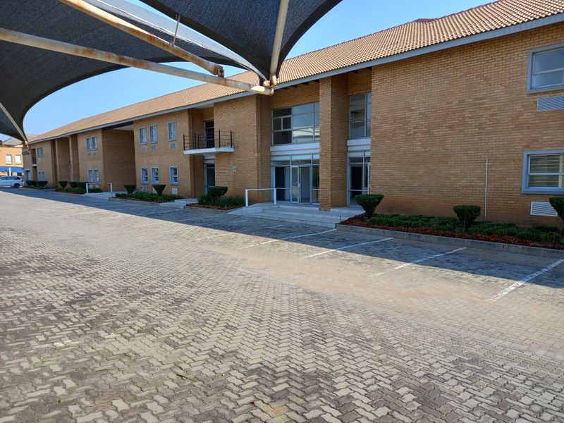 Exterior view of a multi-story office building featuring a brick facade, tiled roof, and a large paved parking area with shade sail structures.