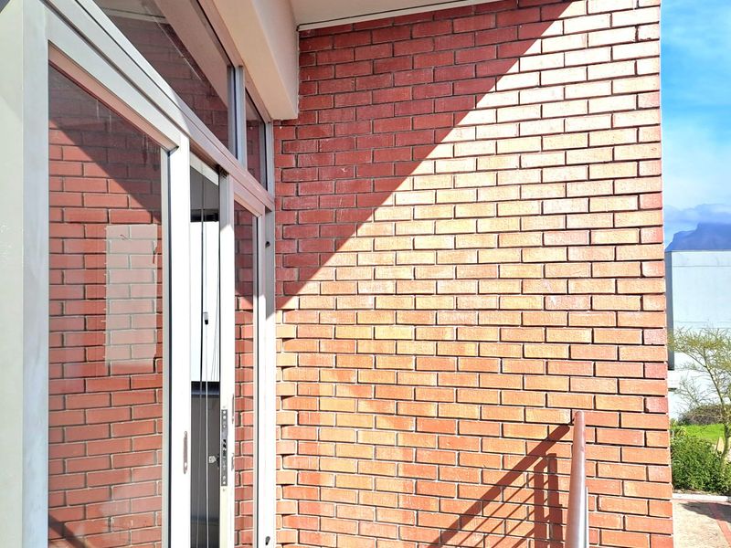 A clean, modern balcony featuring exposed brickwork, aluminium-framed glass sliding doors, and a metal safety railing with tiled flooring.