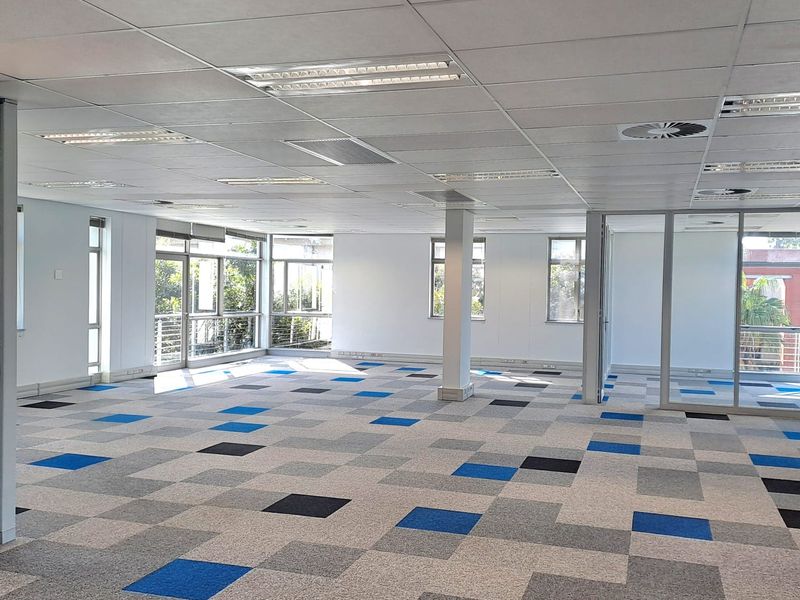 A spacious, open-plan office area featuring modern carpet tiles, suspended ceilings with recessed lighting, and large windows providing ample natural light.