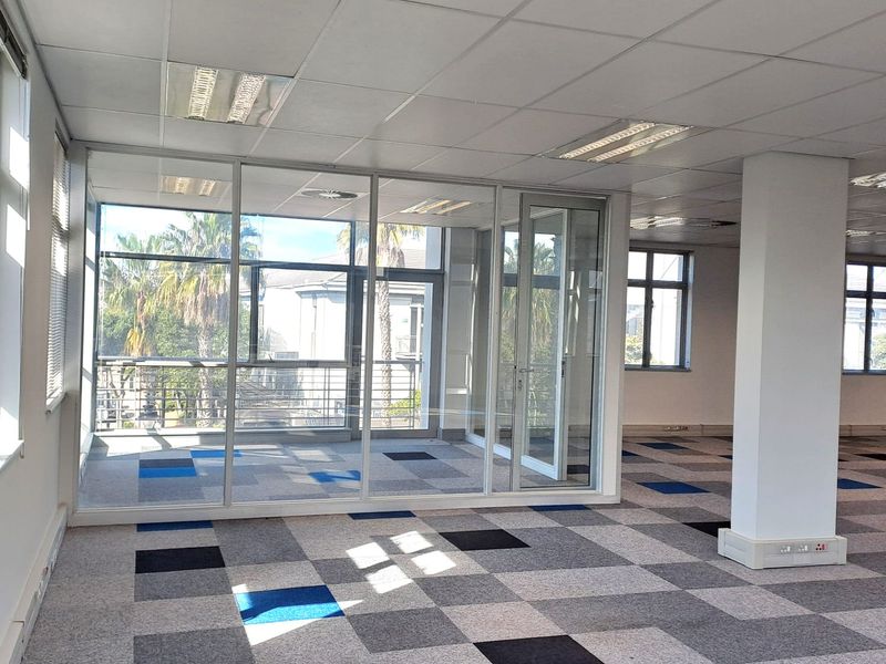 An open-plan office space featuring glass partitioning, suspended ceilings with recessed lighting, and patterned carpet tiles.
