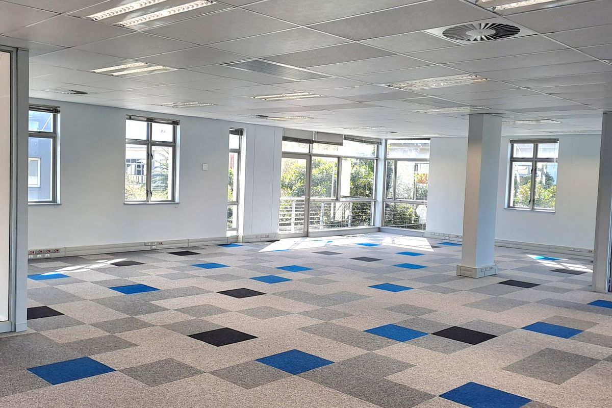 A spacious, open-plan office area featuring a suspended ceiling, recessed lighting, and modern patterned carpet tiles with large windows providing natural light.