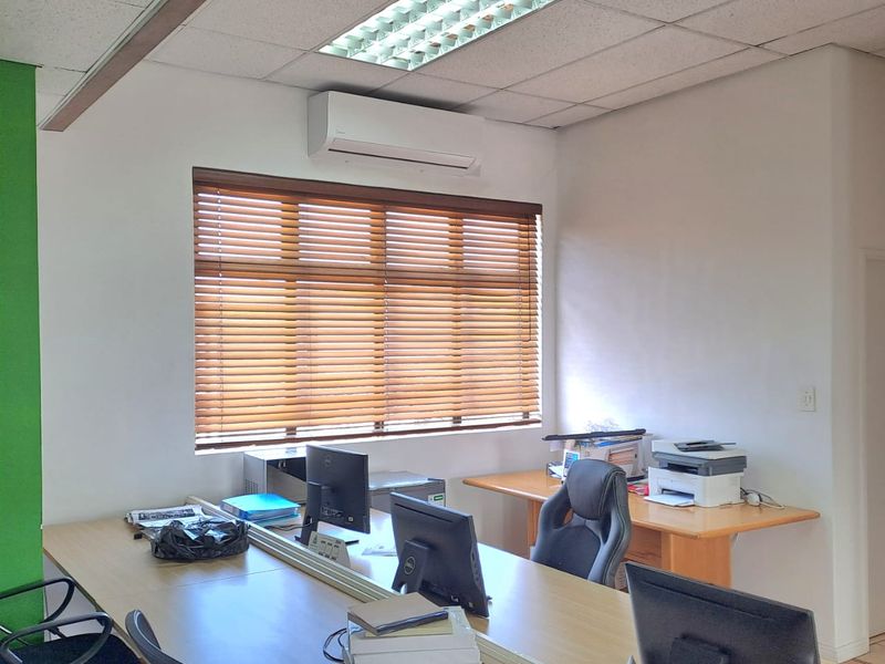 A functional office space featuring a suspended acoustic ceiling, recessed lighting, wall-mounted air conditioning unit, and wooden venetian blinds.