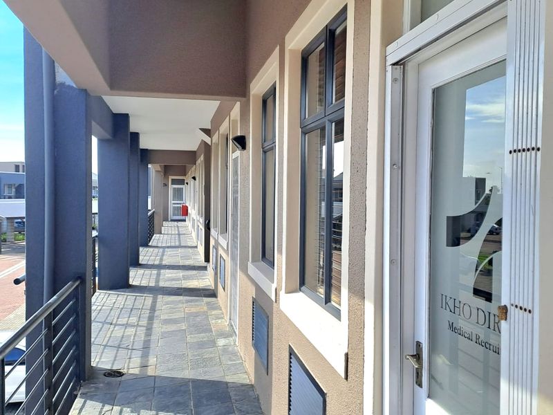 A covered exterior walkway featuring stone tile flooring, structural pillars, and glass-paneled office entrance doors with security gates.
