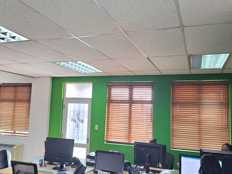 A functional office space featuring suspended acoustic ceiling tiles, recessed lighting, and large windows fitted with wooden venetian blinds.