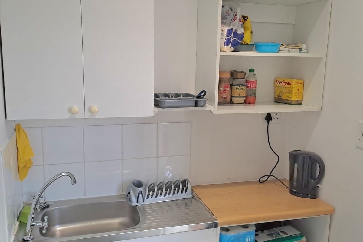 A compact kitchenette area featuring a stainless steel sink, tiled splashback, and basic white cabinetry with open shelving.