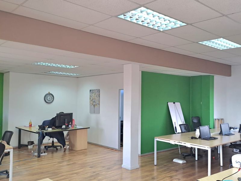 Open-plan office space featuring wood-look laminate flooring, suspended acoustic ceiling tiles, and recessed fluorescent lighting fixtures.
