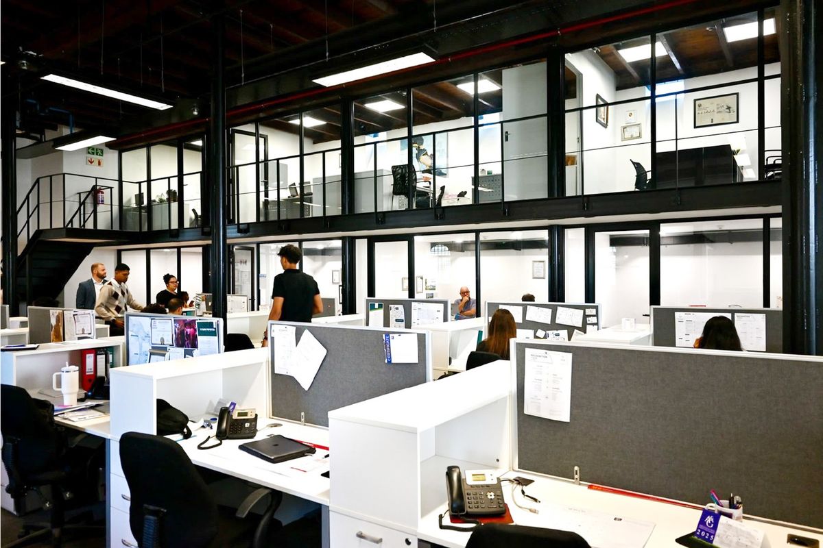 Modern open-plan office space featuring a mezzanine level, industrial exposed ceilings, and glass-partitioned private offices with integrated desk workstations.