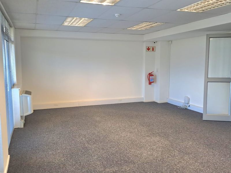 A clean, vacant office space featuring neutral carpeting, suspended ceilings with recessed lighting, and a wall-mounted air conditioning unit.