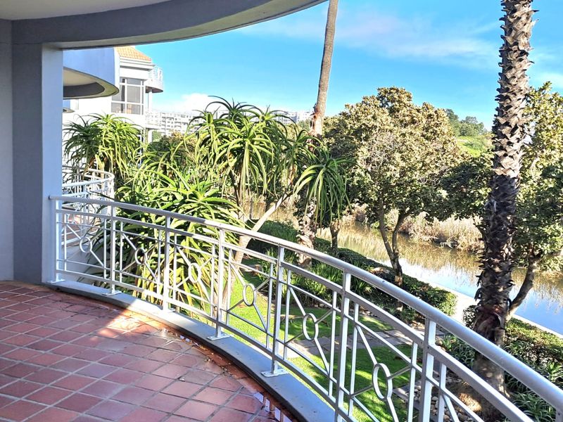 A spacious curved balcony featuring terracotta tiling and a white metal balustrade, overlooking a landscaped garden area with mature trees and a water feature.