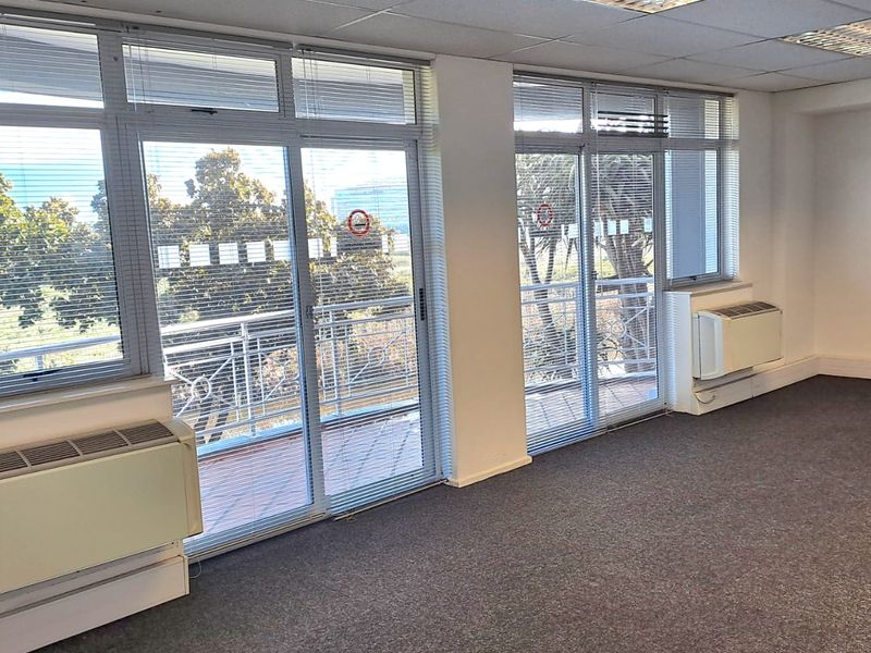 A bright, empty office space featuring carpeted floors, suspended ceilings, wall-mounted air conditioning units, and large sliding glass doors leading to a balcony.