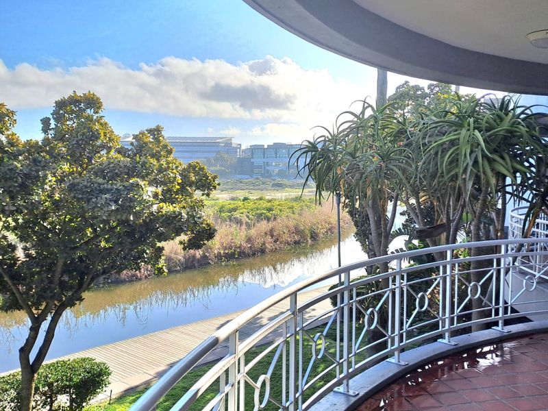 A spacious covered balcony featuring terracotta tiling and a decorative metal balustrade, offering scenic views of the surrounding landscape and water feature.