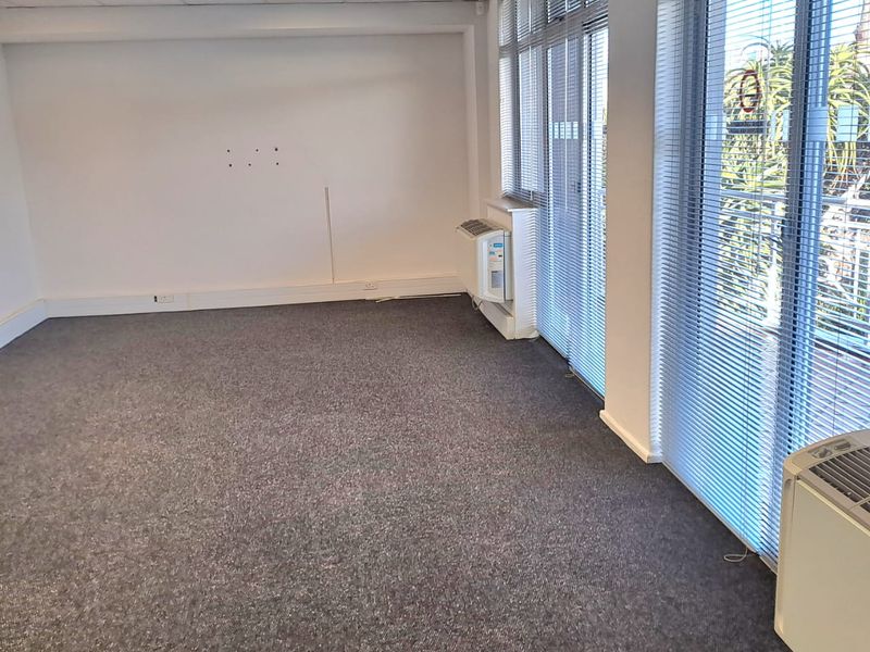 A vacant office space featuring carpeted flooring, suspended ceilings with recessed lighting, air conditioning units, and large windows with venetian blinds.