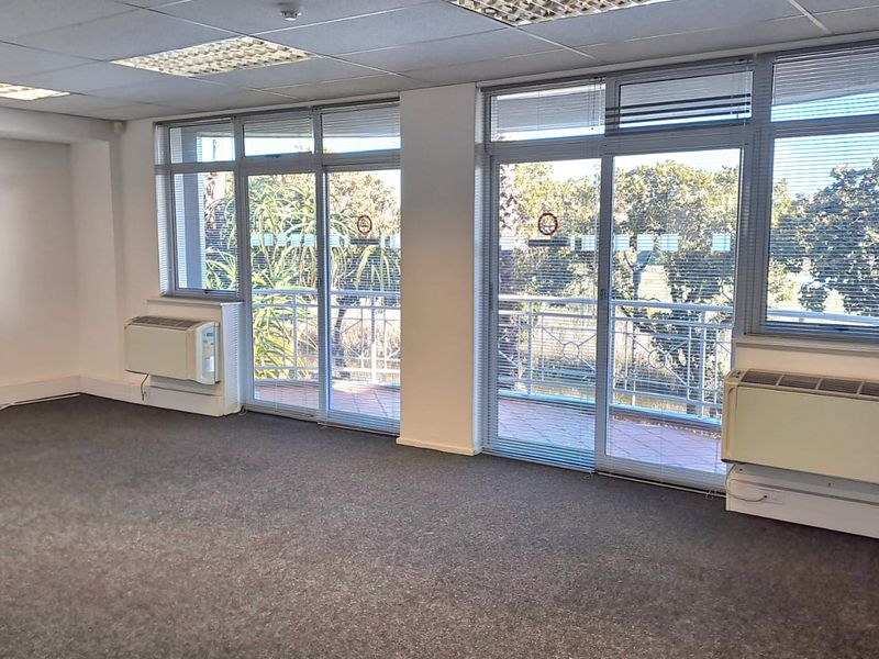 A bright, vacant office space featuring carpeted floors, suspended ceilings, wall-mounted HVAC units, and large sliding glass doors leading to a balcony.