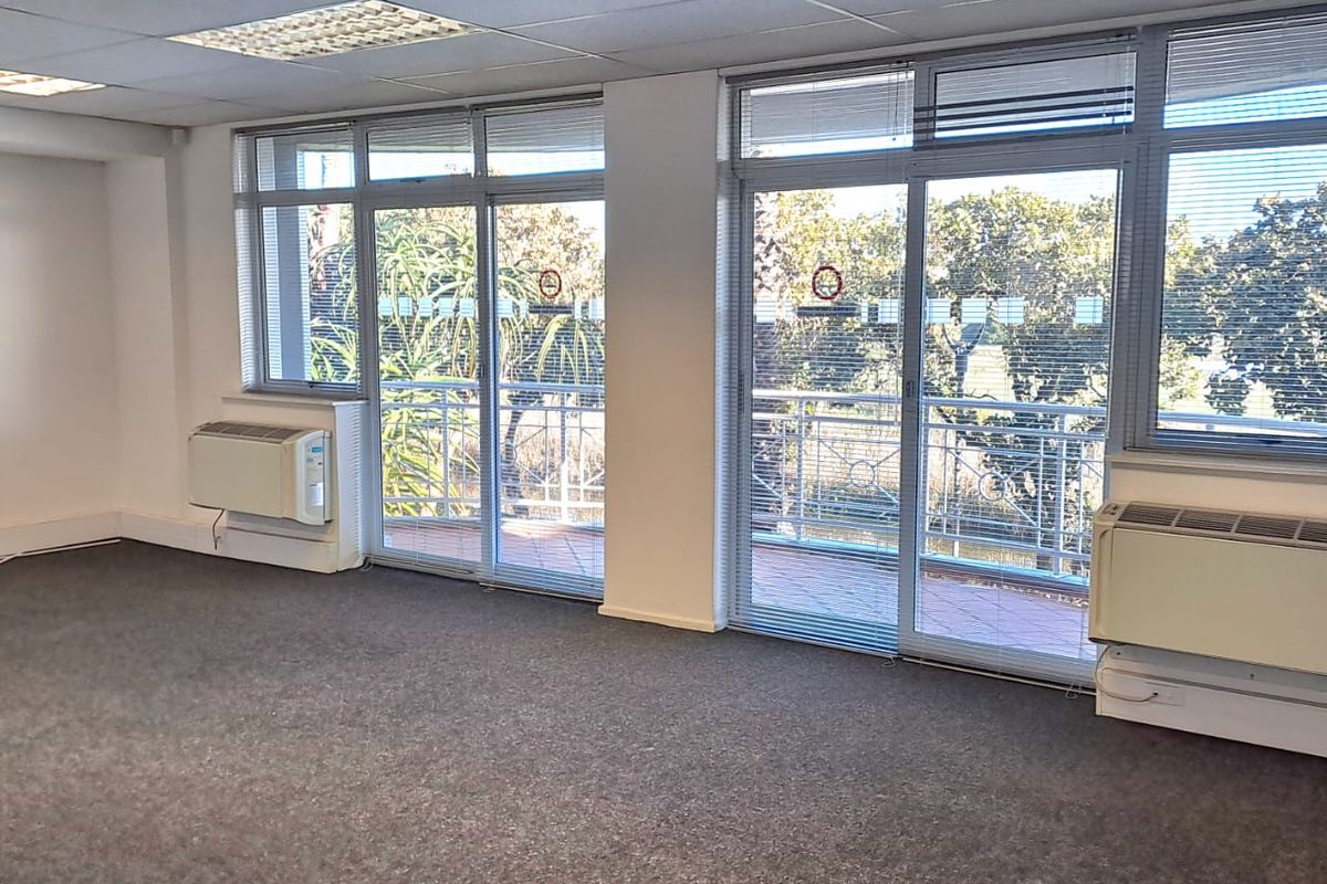 A bright, vacant office space featuring carpeted floors, suspended ceilings, wall-mounted HVAC units, and large sliding glass doors leading to a balcony.
