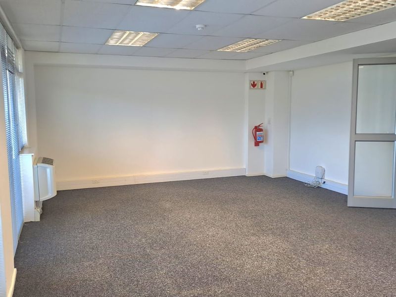 A clean, vacant office space featuring grey carpeting, suspended ceilings with recessed lighting, wall-mounted air conditioning, and a glass-panelled entrance door.