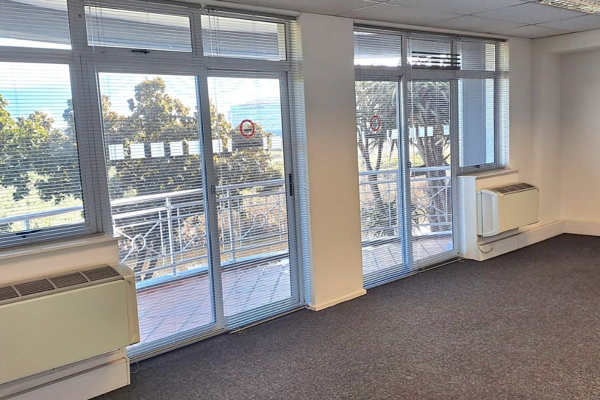A bright, empty office space featuring carpeted floors, suspended ceilings, wall-mounted air conditioning units, and large sliding glass doors leading to a balcony.