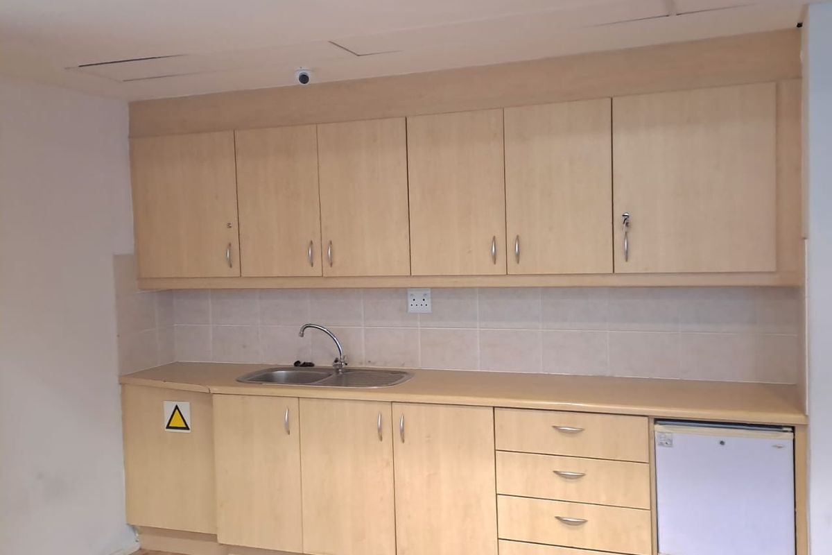 A functional office kitchenette featuring light wood-finish cabinetry, a stainless steel sink, tiled backsplash, and integrated under-counter storage space.