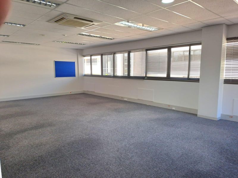 A spacious, empty office area featuring carpeted flooring, a suspended ceiling with recessed lighting, integrated air conditioning, and large windows with fitted blinds.
