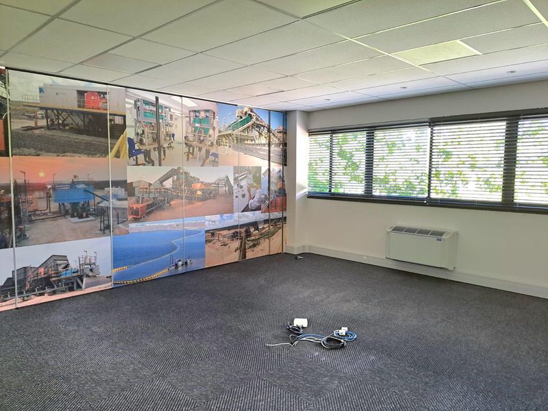 A bright, empty office space featuring carpet tiles, a suspended ceiling, wall-mounted HVAC unit, and a large industrial-themed feature wall mural.