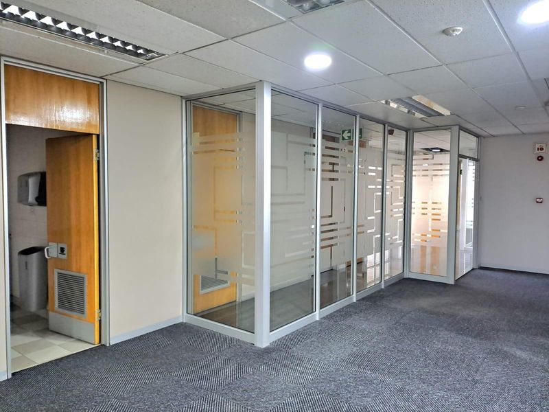A professional office space featuring glass partition walls with frosted detailing, carpeted flooring, and a suspended ceiling with integrated lighting.