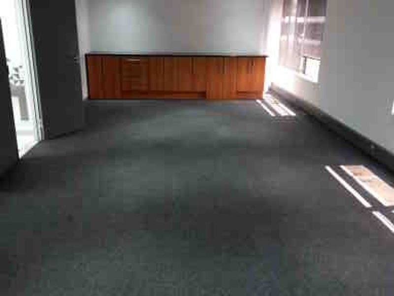 A spacious, empty office area featuring built-in wooden cabinetry, recessed ceiling lighting, a ceiling-mounted air conditioning unit, and dark carpeted flooring.