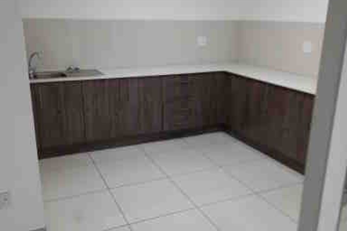 A clean, empty commercial kitchenette featuring dark wood-finish cabinetry, light countertops, a stainless steel sink, and bright tiled flooring under a drop ceiling.