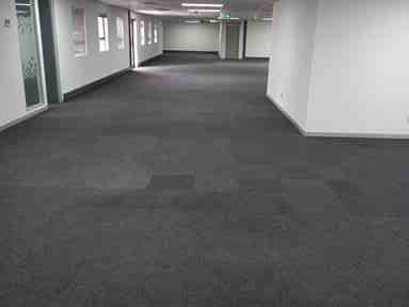 A spacious, empty commercial office area featuring carpeted flooring, a suspended ceiling with recessed lighting, and glass partition walls.