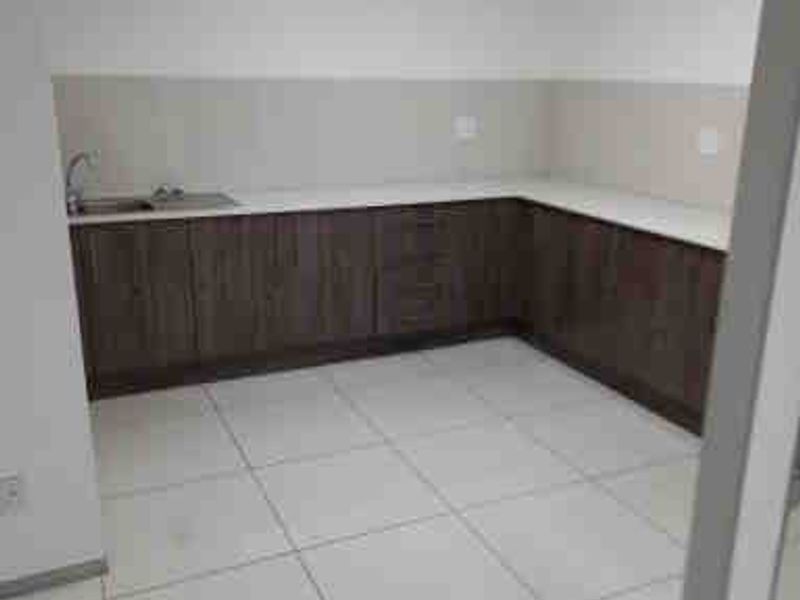 A clean, empty commercial kitchenette featuring dark wood-finish cabinetry, light countertops, a stainless steel sink, and bright tiled flooring under a drop ceiling.