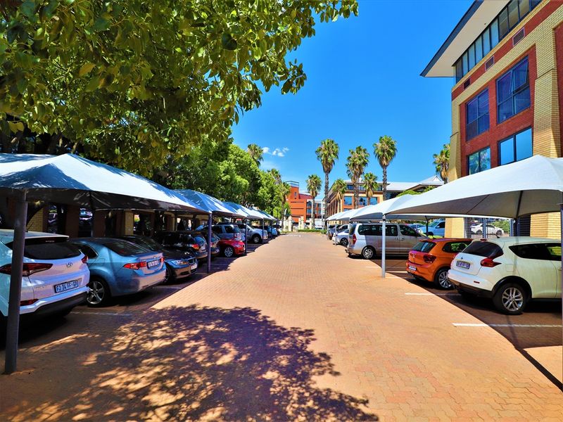 A well-maintained office parking area featuring paved surfaces and structured shade cloth carports, situated alongside a multi-story brick office building.