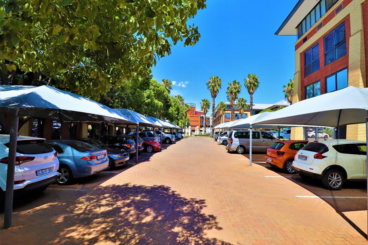 A well-maintained office parking area featuring paved surfaces and structured shade cloth carports, situated alongside a multi-story brick office building.