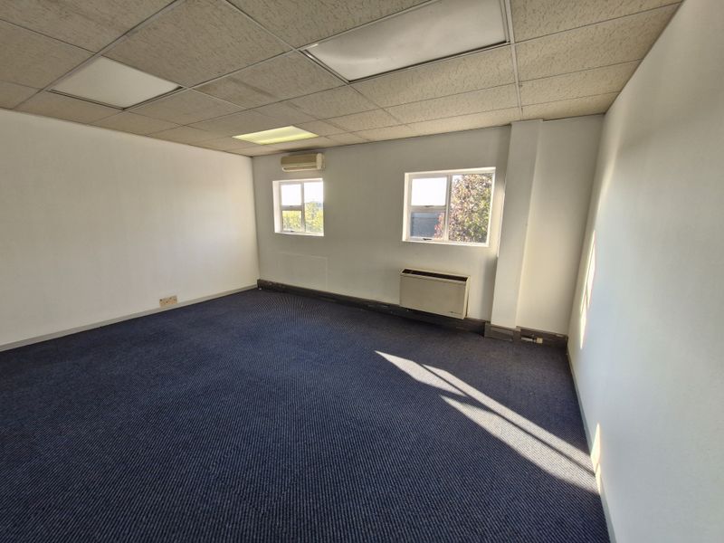 An empty office space featuring a suspended ceiling, wall-mounted air conditioning, baseboard heating, and carpet flooring with natural light from two windows.