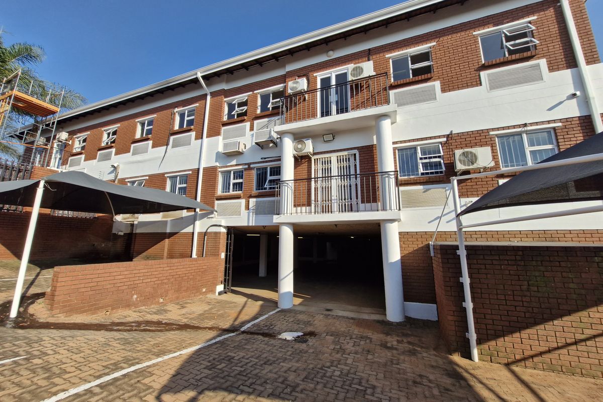 Rear view of a multi-story office building featuring brick facade, balconies with metal railings, external air conditioning units, and covered parking area.