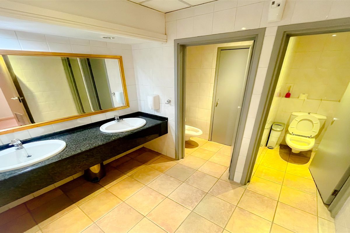 A functional office restroom featuring a dual-basin vanity with granite countertop, tiled flooring, and separate enclosed toilet cubicles.