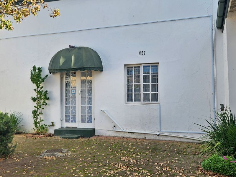 Rear exterior view of the commercial property featuring a paved courtyard, white plastered walls, a green awning over french doors, and a lattice window.