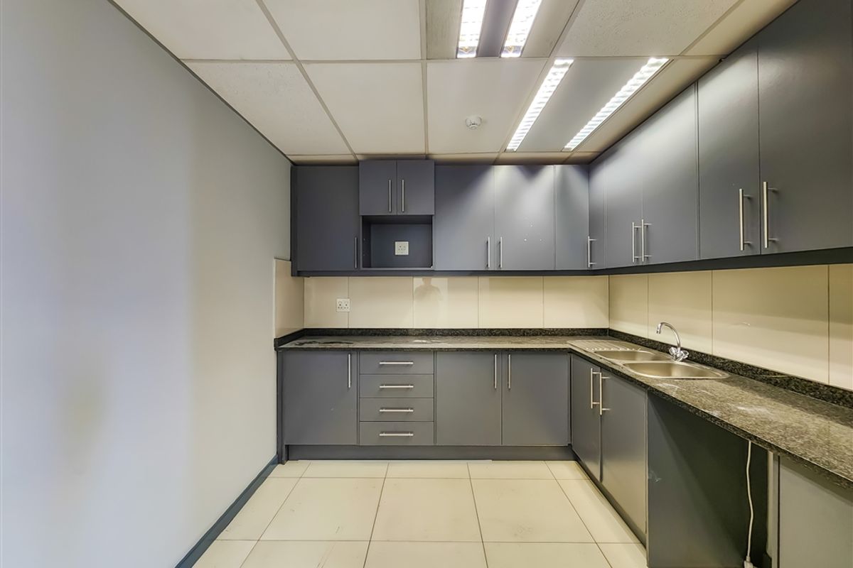 A functional office kitchenette featuring dark grey cabinetry, granite countertops, a stainless steel sink, and integrated fluorescent lighting.