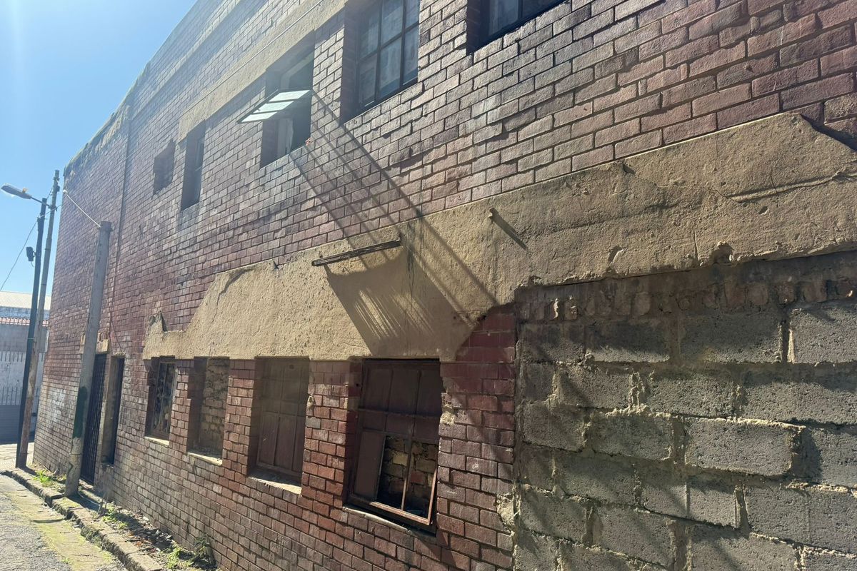 Exterior view of an industrial brick building showing significant wear to the facade, damaged window frames, and crumbling concrete structural elements.