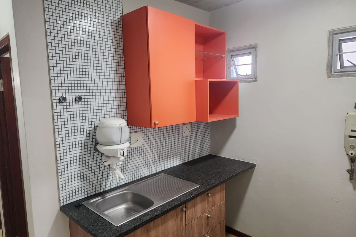 A functional kitchenette area featuring wood-look cabinetry, a dark countertop, stainless steel sink, and a mosaic tiled splashback.