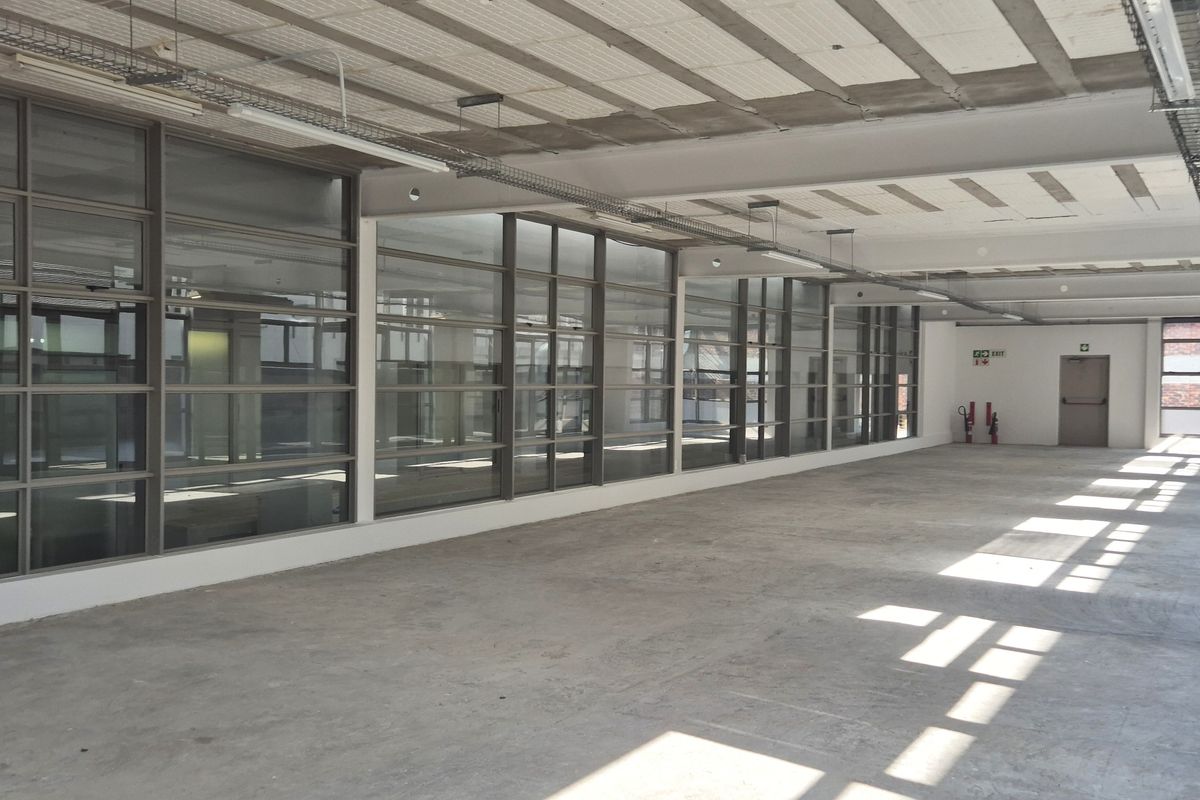 Spacious open-plan office area featuring large glass partitions, exposed concrete ceilings with cable management systems, and ample natural light.