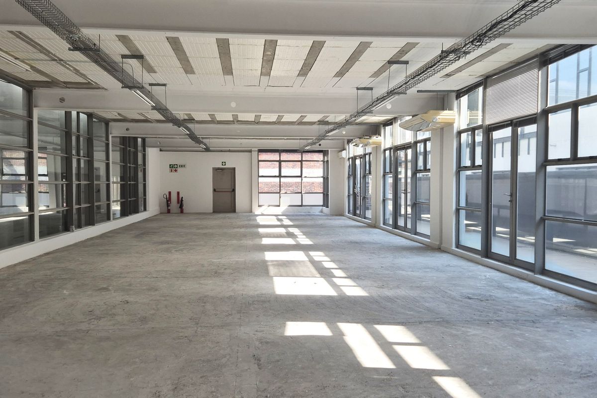 Spacious open-plan office area featuring large windows, exposed industrial-style ceiling, and concrete flooring, offering a versatile workspace with abundant natural light.