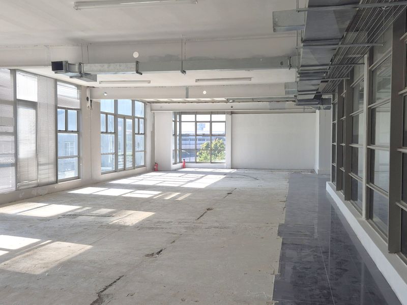 A spacious, open-plan office area featuring large windows, exposed industrial-style ceiling ductwork, and unfinished concrete flooring, offering a blank canvas for customization.