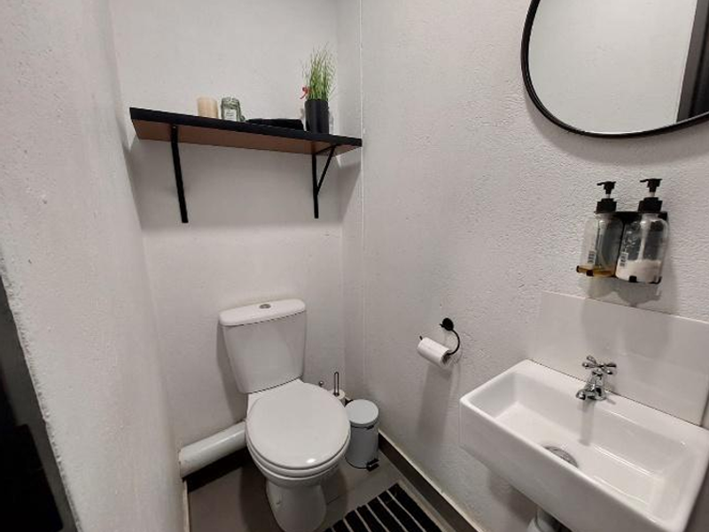 A compact, functional office restroom featuring a wall-mounted basin, modern toilet, and a floating shelf for storage.